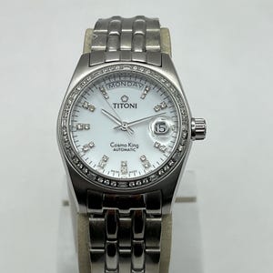 Puede incluir: Un reloj de pulsera automático Titoni Cosmo King plateado con esfera blanca, detalles de diamantes y visualización de la fecha. El reloj tiene una correa plateada y se muestra sobre un soporte de tono neutro. La esfera muestra el día de la semana como "Lunes".