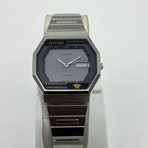 Puede incluir: Un reloj automático Citizen plateado con esfera gris y visualización de día y fecha. El reloj tiene una caja rectangular con una correa plateada a juego. La esfera muestra el día como "FRI 2".
