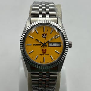 Puede incluir: Un reloj de pulsera Rado de acero inoxidable con esfera amarilla. El reloj presenta un bisel estriado, una pantalla de fecha y una pulsera plateada. La esfera tiene el texto "RADO" y "Purple Horse".