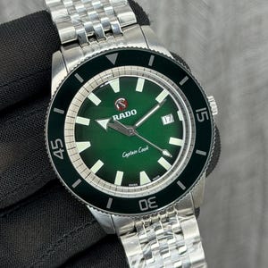 Può includere: Un orologio Rado Captain Cook in acciaio inossidabile con quadrante verde e datario a ore 3. L'orologio ha una lunetta nera e un bracciale color argento. Il quadrante mostra il logo Rado e le parole "Captain Cook".