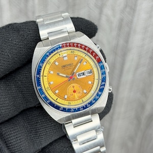 Puede incluir: Un reloj automático Seiko Chronograph vintage con una correa de acero inoxidable plateado. La esfera del reloj es dorada con detalles en azul, rojo y amarillo. Se muestra el día y la fecha. Un reloj clásico.