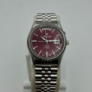 Peut inclure: Une montre-bracelet automatique Ricoh argentée avec un cadran bordeaux. La montre présente une lunette cannelée, un affichage de la date et un bracelet en acier inoxydable. Le cadran affiche "Wednesday", "Ricoh", "Automatic" et "21 Jewels".