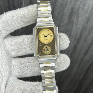 May include: A vintage wristwatch with a rectangular gold-toned face and a silver and gold-toned metal band. The watch face features a date display and the text "delil Syrea". The watch is held by a gloved hand.