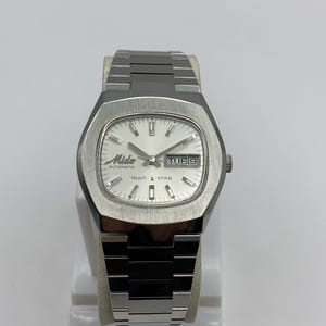 Puede incluir: Un reloj automático Mido Multi Star plateado con esfera blanca y visualización de día y fecha. El reloj tiene una caja rectangular y una correa de acero inoxidable. El reloj se muestra sobre un soporte blanco.