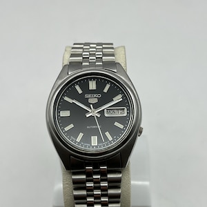 May include: A stainless steel Seiko automatic watch with a black dial. The watch features silver hour markers, hands, and a day-date display. The watch is displayed on a white stand. The watch band is made of stainless steel.