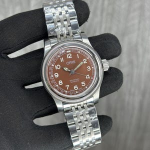 Puede incluir: Un reloj de pulsera automático Oris Big Crown con esfera marrón, manecillas plateadas y números arábigos. El reloj cuenta con una aguja de fecha, un bisel estriado y una pulsera de eslabones de acero inoxidable con película protectora.