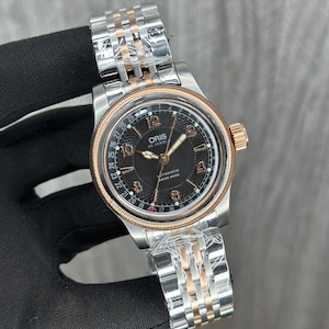 Puede incluir: Un reloj de pulsera de acero inoxidable con esfera negra y correa bicolor plateada y oro rosa. La esfera del reloj muestra la marca "ORIS" y el texto "AUTOMATIC SWISS MADE". El reloj tiene una visualización de la fecha alrededor de la esfera.