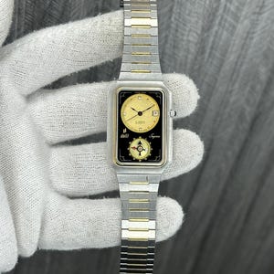 May include: A rectangular wristwatch with a gold and silver strap. The watch face is gold with black detailing and features the text "doll" and "Signe". The watch has a date display and a compass design.