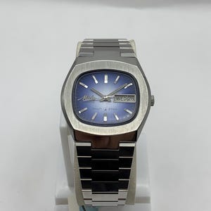Puede incluir: Un reloj Mido Multi Star plateado con esfera azul y agujas plateadas. El reloj tiene una caja rectangular y una correa de metal. El día y la fecha se muestran a las 3 en punto. El reloj se exhibe sobre un soporte blanco.
