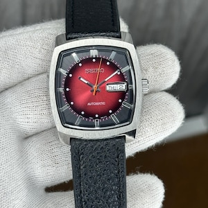 Puede incluir: Un reloj automático Seiko de acero inoxidable con una correa de cuero negro. La esfera del reloj es de color rojo intenso con marcadores de hora y agujas plateadas. El día y la fecha se muestran en una pequeña ventana. El reloj es rectangular.