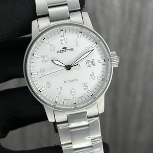 May include: A silver wristwatch with a white face and a matching metal strap. The watch face displays black numerals, hands, and the word "FORTIS". The watch also shows the word "AUTOMATIC" and a date window.