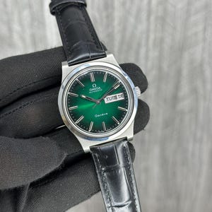 Pode incluir: Um relógio de pulso automático Omega Genève com caixa redonda prateada e mostrador verde esmeralda com efeito de raios de sol. O relógio inclui índices de bastão prateados, uma janela de dia e data às três horas e uma pulseira de couro preta.
