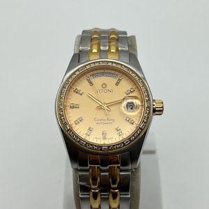 Puede incluir: Un reloj de pulsera automático Titoni Cosmo King con esfera dorada y correa plateada y dorada. La esfera muestra "Thursday" y tiene una ventana de fecha. El bisel está incrustado con pequeñas piedras transparentes.