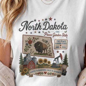 May include: A white t-shirt featuring a graphic print of North Dakota, The Peace Garden State. The design includes a bison, a red barn, sunflowers, an oil pump, and text reading North Dakota, The Peace Garden State, 1889-2025, and Rooted in Ranching, Rich in Resources.