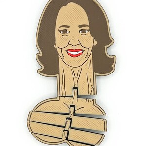 May include: A wooden ornament featuring a smiling face with red lipstick and brown hair. The ornament has a unique design with a cutout section below the face. The ornament is light brown and dark brown.