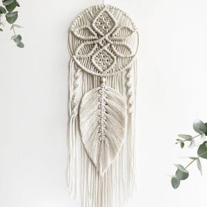 May include: A cream-coloured macrame wall hanging. It features a circular design with a geometric pattern at the top and a large, textured feather shape below. The piece is suspended from a gold-coloured metal ring and includes hanging strands.
