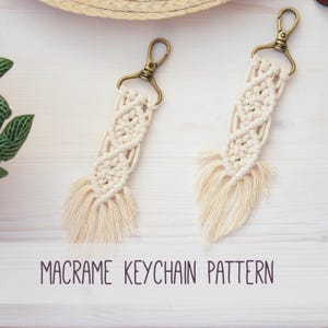 Pode incluir: Dois chaveiros de macramê creme com fechos e franjas bronze. Exibidos em uma superfície de madeira branca. O texto "MACRAME KEYCHAIN PATTERN" está impresso em roxo escuro.