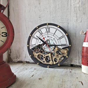 Può includere: Un orologio rotondo con cornice nera e quadrante bianco con numeri romani e la scritta "Mount Everest". L'orologio ha ingranaggi visibili e dettagli dorati. L'orologio ha un diametro di circa 25 cm.