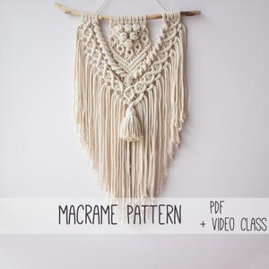 May include: Cream-coloured macrame wall hanging with a geometric design, suspended from a wooden branch. The piece features intricate knotting and a central tassel. Text at the bottom reads "MACRAME PATTERN PDF + VIDEO CLASS."