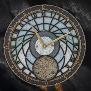 May include: A round, decorative clock with a dark blue and gold frame. The clock face features Roman numerals, gold hands, and a detailed, geometric design with light blue and white accents. The clock has a vintage aesthetic.