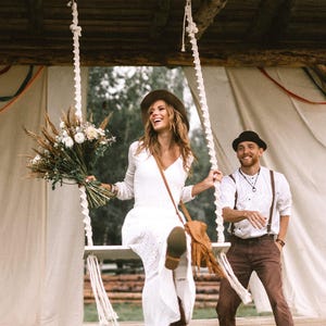 Op de afbeelding: Een vrouw in een witte jurk en bruine hoed zit op een schommel en houdt een boeket bloemen vast. Een man in een wit overhemd en bruine broek staat in de buurt. De scène speelt zich buiten af met een houten constructie en een tent op de achtergrond.