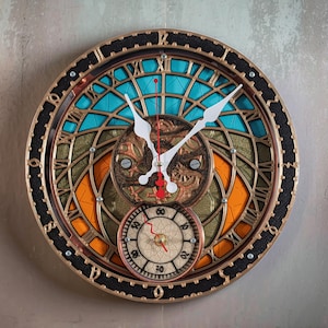 May include: A round, multi-layered wall clock with a complex design. The clock features a gold-colored frame, black accents, and a combination of teal, orange, and gold-colored geometric patterns. White clock hands indicate the time. A smaller clock is at the bottom.