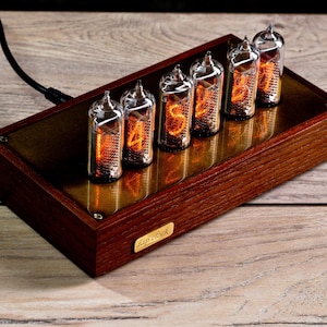 Könnte beinhalten: Eine Digitaluhr mit sechs Glasröhren, die leuchtende orangefarbene Zahlen anzeigen. Die Uhr befindet sich in einem rechteckigen Holzrahmen mit einer messingfarbenen Metallplatte. Die Uhr ist mit einem schwarzen Kabel angeschlossen.