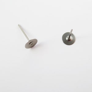 May include: Two silver-colored earring posts with flat, circular backs and thin, straight posts. The posts are on a white background, with one post lying down and the other standing upright. The earring posts are simple and minimalist.