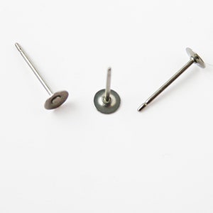 May include: Three silver-colored stud earrings with a flat, circular base and a thin, straight post. The earrings are displayed on a white background, with the posts angled in different directions.