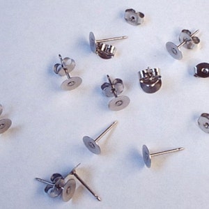 May include: A collection of silver-colored earring posts and backs scattered on a white surface. The earring posts have a flat, round disc for attaching the earring, and a straight post for insertion. The backs are butterfly style.