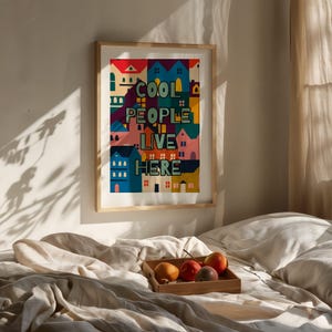 Può includere: Una stampa artistica incorniciata con la scritta "COOL PEOPLE LIVE HERE" in un design colorato e stilizzato di case. L'opera presenta una varietà di colori, tra cui blu, rosso e giallo. La stampa è esposta su una parete sopra un letto.