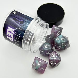 May include: A clear plastic container with a black lid, containing several polyhedral dice. The dice are a mix of purple, teal, and silver, with white numbering. The container has a label that reads "GHOST KEY".