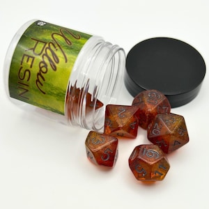 May include: A clear plastic jar with a black lid and several orange and gold resin dice. The jar has a green and yellow label with the text "Willow Resin". The dice are scattered on a white surface.