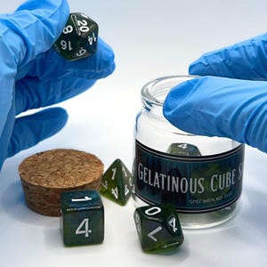 May include: A set of dark green translucent polyhedral dice with white numbering. One twenty-sided die is held up, while others sit near a clear glass jar with a cork stopper. The jar label reads Gelatinous Cube Specimen No. 003 in white text.