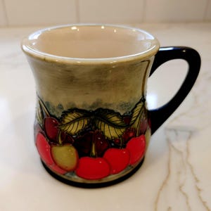 May include: A ceramic mug with a black handle and a cream-colored interior. The exterior features a painted design of red cherries and green leaves against a muted background. The mug has a slightly flared shape.