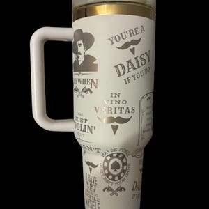 May include: A white insulated tumbler with a handle, featuring a clear lid and gold trim. The tumbler is adorned with various text and graphics, including a cowboy, mustaches, and phrases like "You're a Daisy" and "In Vino Veritas."