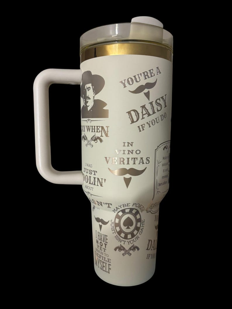 May include: A white insulated tumbler with a handle, featuring a clear lid and gold trim. The tumbler is adorned with various text and graphics, including a cowboy, mustaches, and phrases like "You're a Daisy" and "In Vino Veritas."