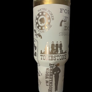 May include: A white tumbler with a gold rim and lid, featuring a design with the word "Tombstone" and other text and graphics related to the Wild West. The design includes a poker chip, a gun, and the phrase "I'm your Huckleberry."