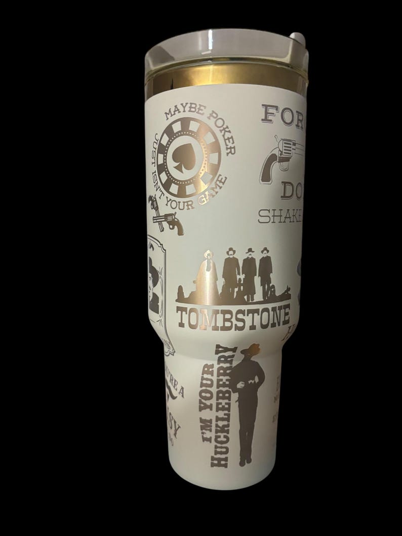 May include: A white tumbler with a gold rim and lid, featuring a design with the word "Tombstone" and other text and graphics related to the Wild West. The design includes a poker chip, a gun, and the phrase "I'm your Huckleberry."