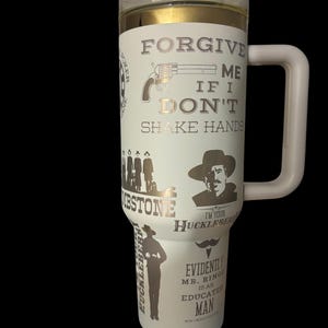 May include: White travel mug with a clear lid and a large handle. The mug features gold and silver graphics and text, including a revolver, the words "FORGIVE ME IF I DON'T SHAKE HANDS," and other Western-themed imagery.