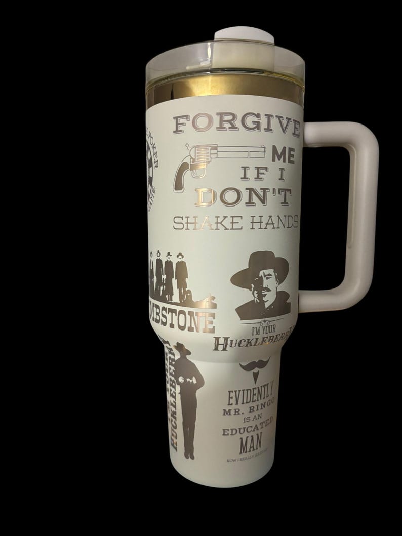 May include: White travel mug with a clear lid and a large handle. The mug features gold and silver graphics and text, including a revolver, the words "FORGIVE ME IF I DON'T SHAKE HANDS," and other Western-themed imagery.