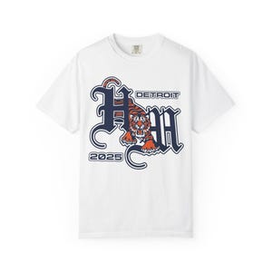 May include: White t-shirt featuring a tiger graphic in orange and black, with the word "DETROIT" above and the year "2025" below. The design is in a vintage, gothic style.