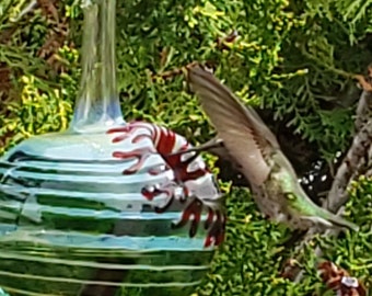 Può includere: Un alimentatore per colibrì in vetro con un design a strisce verde, bianco e rosso. L'alimentatore ha un design a fiore rosso e bianco sul lato. Un colibrì è appollaiato sull'alimentatore.