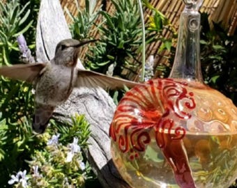 May include: A hummingbird flies towards a decorative glass bird feeder. The feeder has a round, clear glass body with red and orange swirl designs. The background features green plants and a wooden structure.