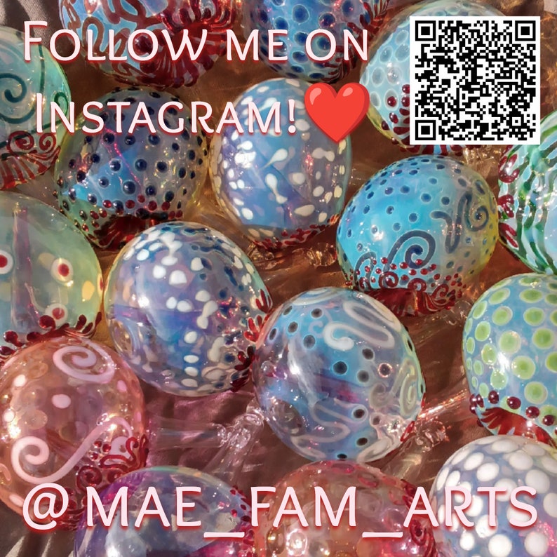 May include: A collection of colorful glass ornaments with intricate designs, including swirls, dots, and white accents. The ornaments are arranged on a pink surface with a QR code and the text "FOLLOW ME ON INSTAGRAM!" in the background.