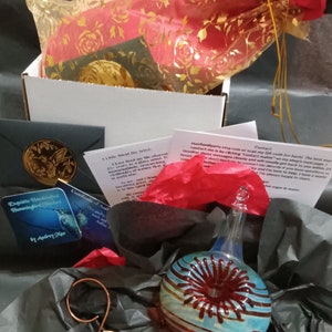 May include: A gift box filled with tissue paper, a small card, and a glass ornament. The ornament is a blue and red glass sphere with a red swirl design. The card reads "Exquisite Handcrafted Hummingbird by Audrey Kaa".