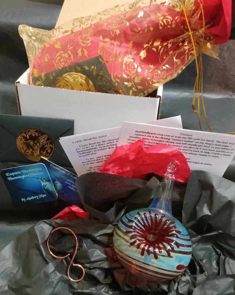 May include: A gift box filled with tissue paper, a small card, and a glass ornament. The ornament is a blue and red glass sphere with a red swirl design. The card reads "Exquisite Handcrafted Hummingbird by Audrey Kaa".