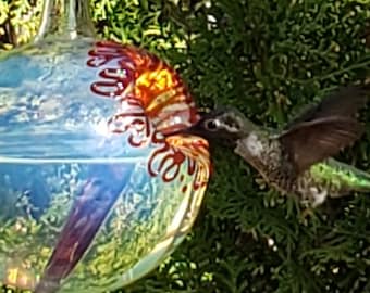 May include: A hummingbird drinks from a glass bird feeder. The feeder is round with a blue and clear glass body and red and orange decorative accents. The bird has brown and green feathers.