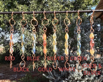 May include: Eight glass and copper suncatchers hanging from a metal rod. The suncatchers are in various colours, including red, blue, silver, green, gold, and black. The suncatchers are made of glass beads and copper wire.