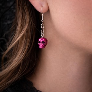 May include: A single pink skull earring with a silver chain and hook. The skull is a vibrant magenta color and has detailed facial features. The earring hangs from a silver hook.